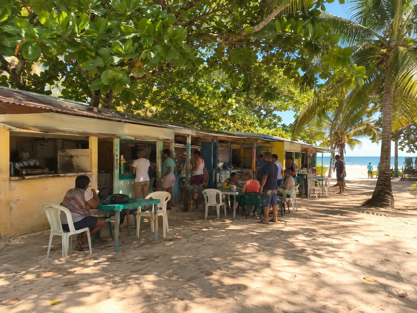 Una fila de kioscos de comida de madera contrachapada descolorida bajo árboles de uva de mar, mesas de plástico colocadas frente a ellos